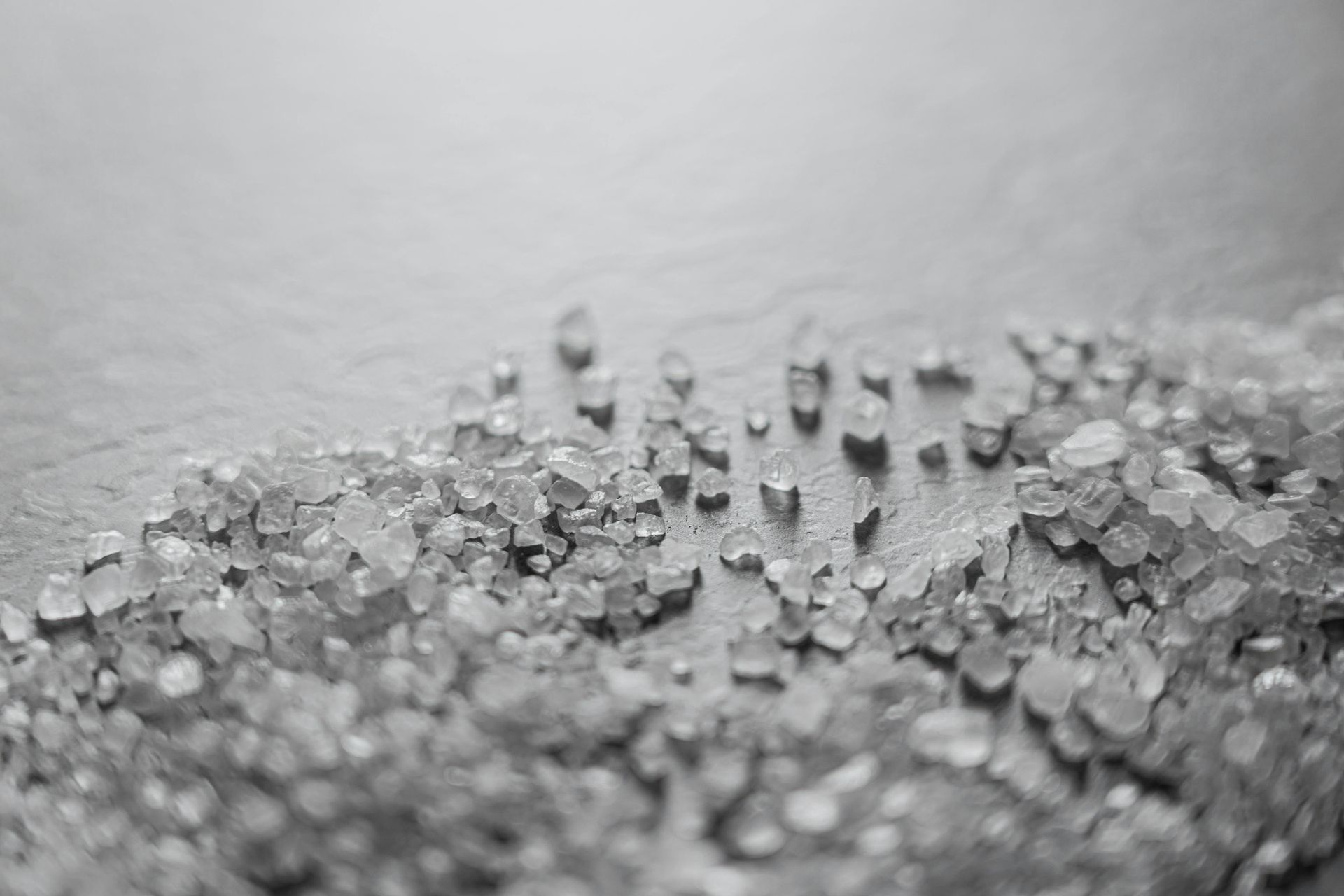 A close-up, black-and-white image of scattered coarse sea salt crystals on a dark, textured surface.