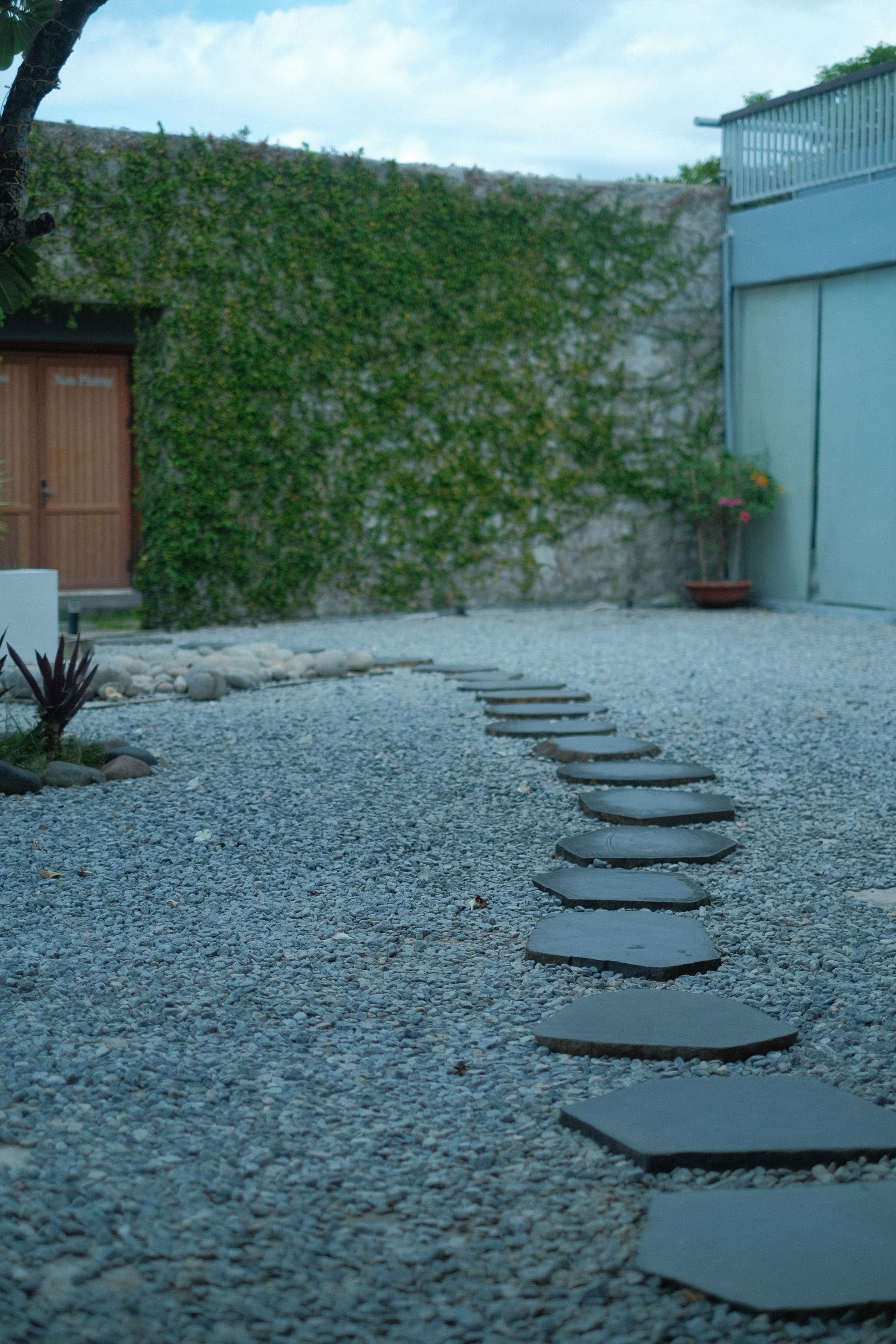 Gravel courtyard with stepping stones leading to a vine-covered wall and blue building.