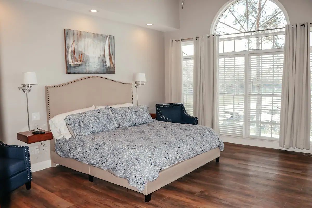 Bright bedroom with a beige upholstered bed, blue bedding, wood floor, and large arched windows with blinds