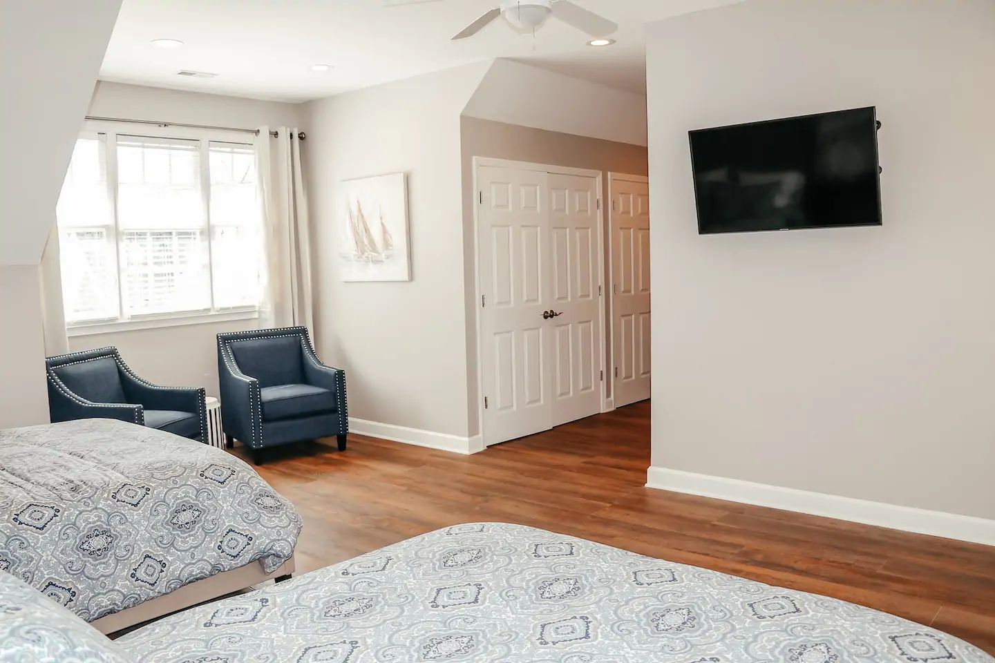 Bright bedroom with two blue chairs, a wall-mounted TV, and a double bed by a window.