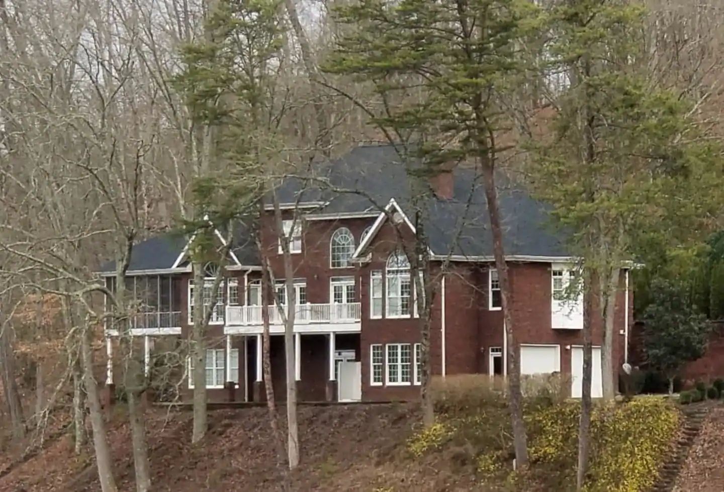 Large red-brick house with white trim on a wooded hillside in autumn