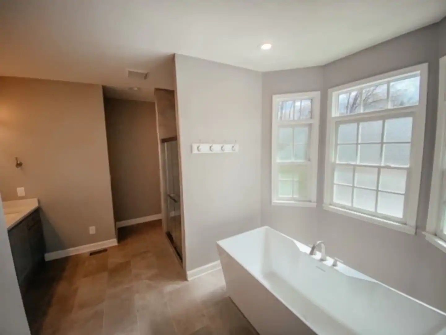 Bright bathroom with white freestanding tub, double windows, beige walls, and walk-in closet opening