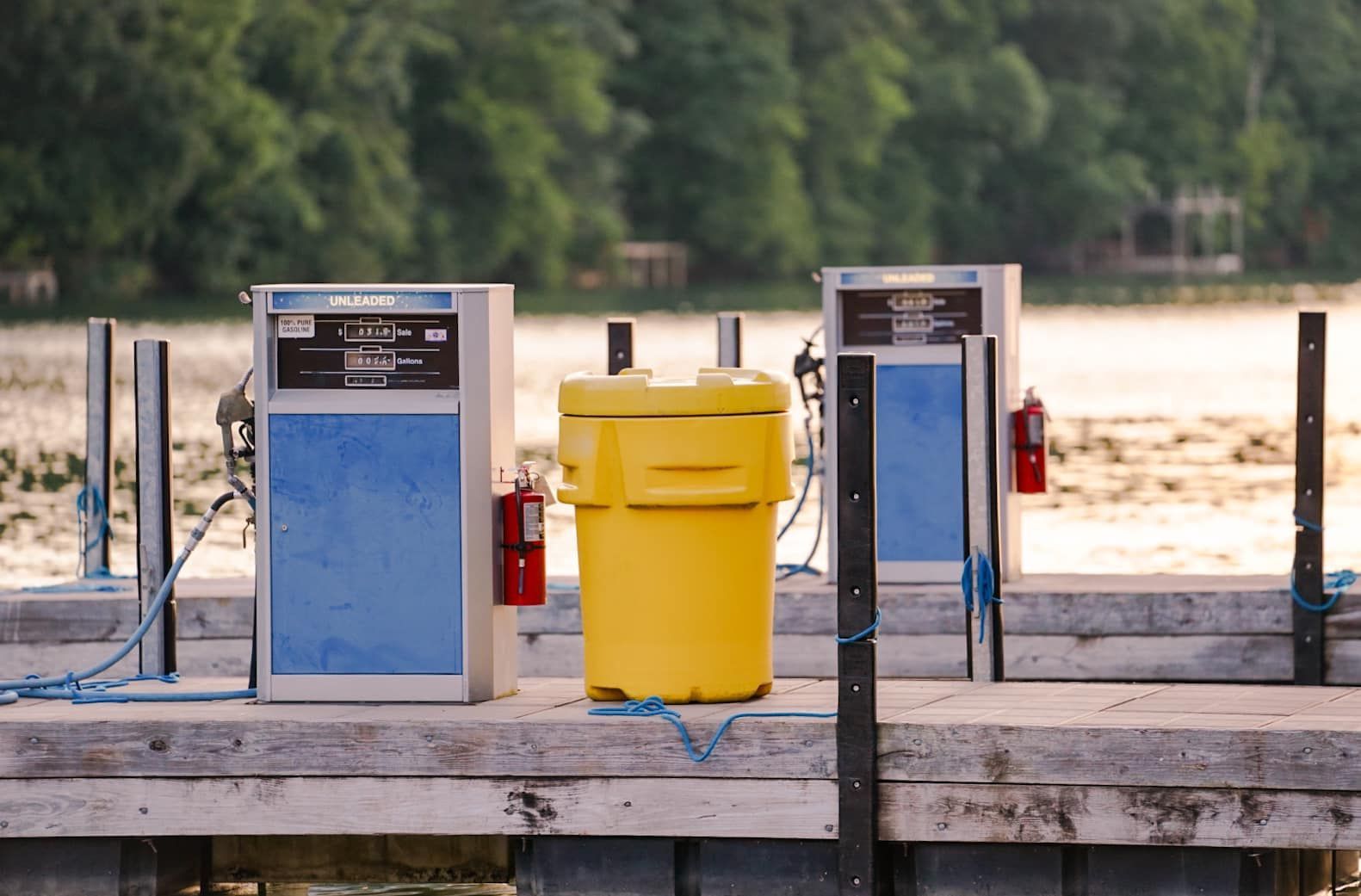 Blue and yellow trash bins on a wooden dock by a lake, with trees in the background
