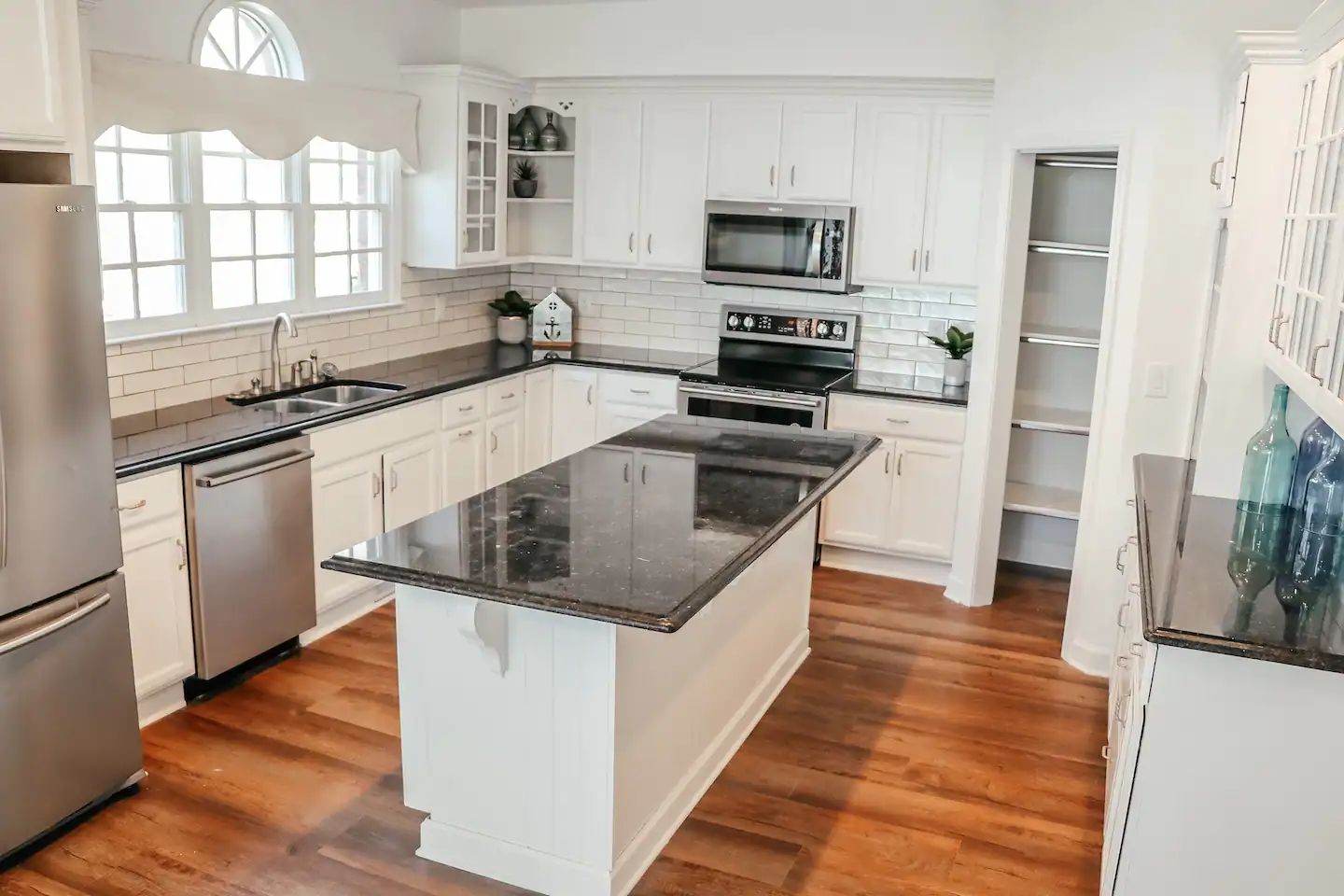 Bright white kitchen with stainless appliances, dark island countertop, and warm wood floors