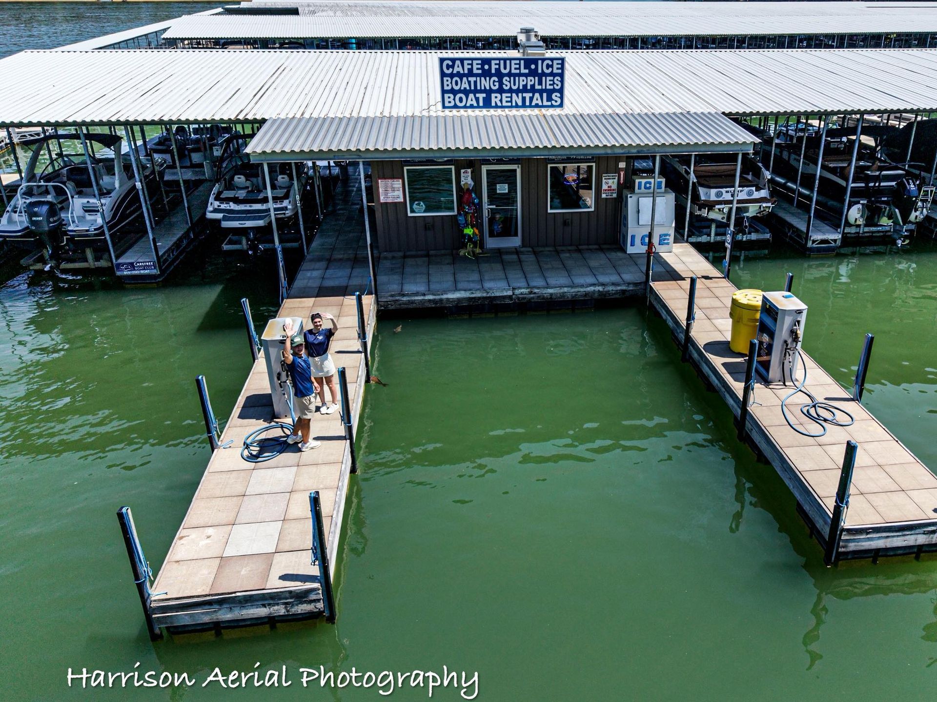 Marina dock with covered boat slips and two people on a wooden walkway beside green water