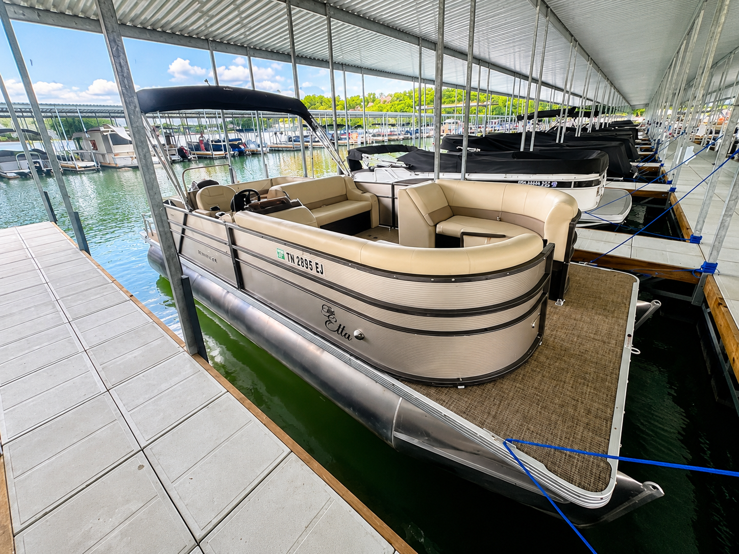 Rent This Boat! Pontoon boat with cream seats docked at a marina under a covered canopy, beside calm water.