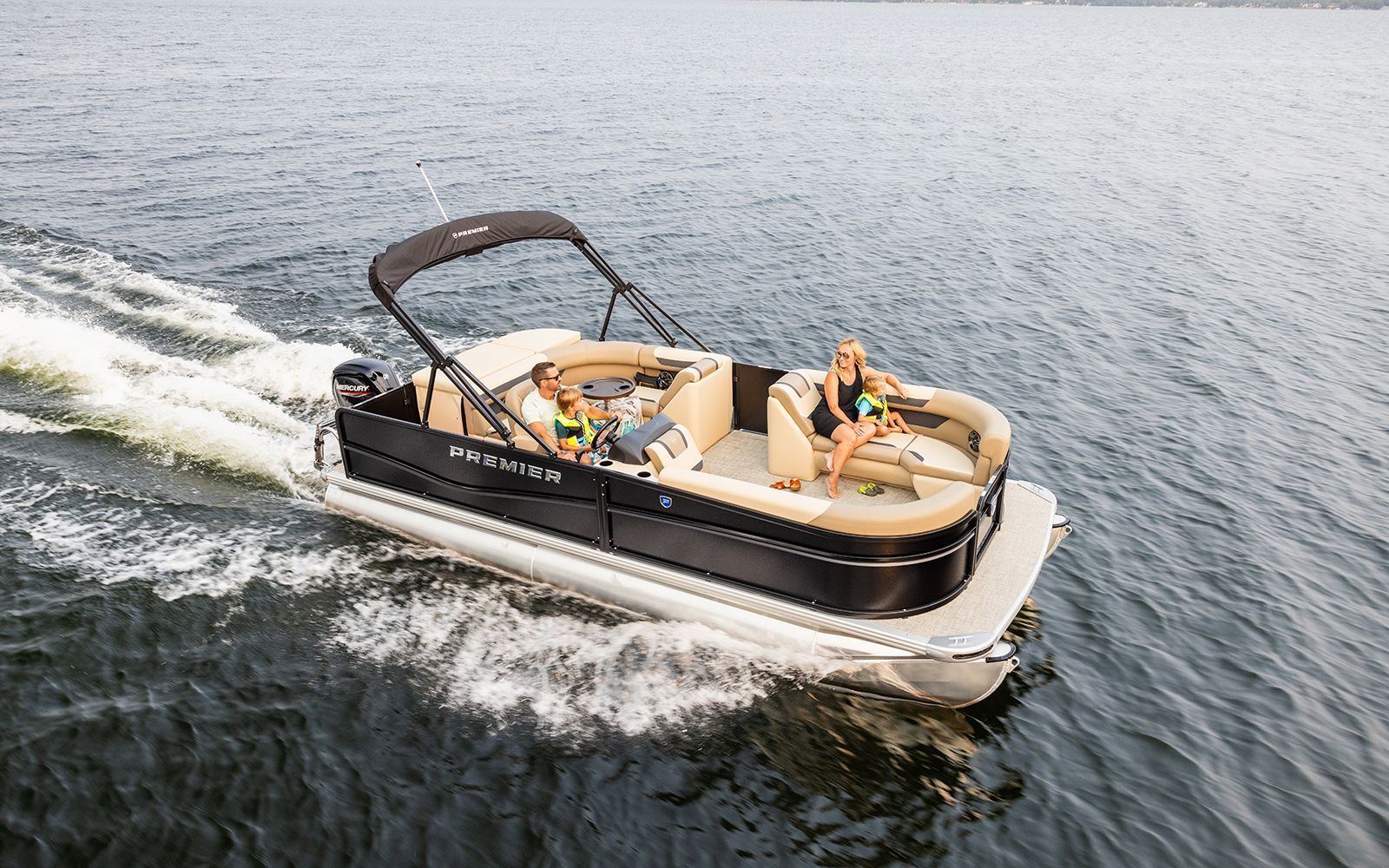 Rent This Boat! Pontoon boat cruising on open water with two people lounging on tan seats under a black canopy.