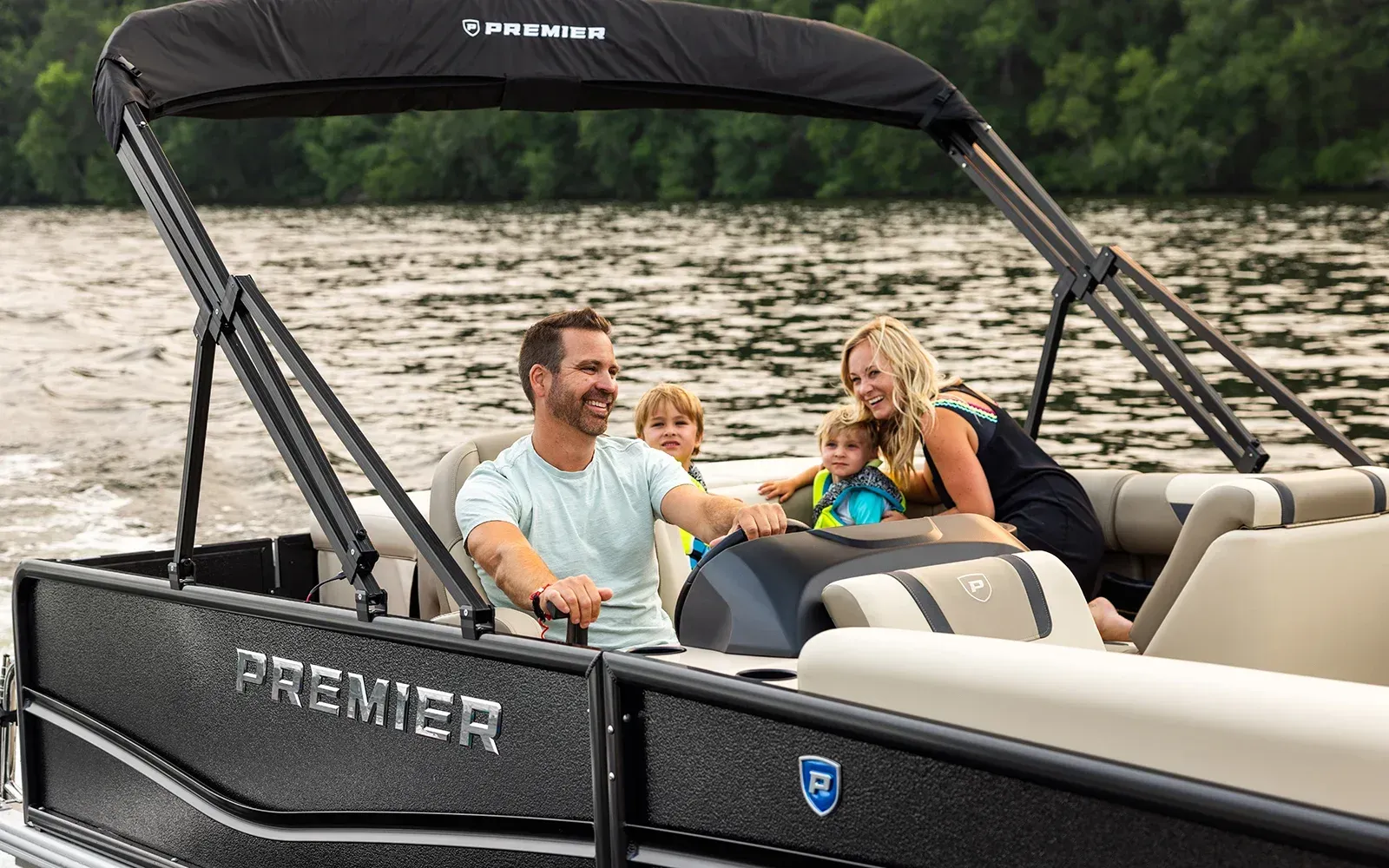 Family riding in a black pontoon boat on a calm lake, smiling under a canopy