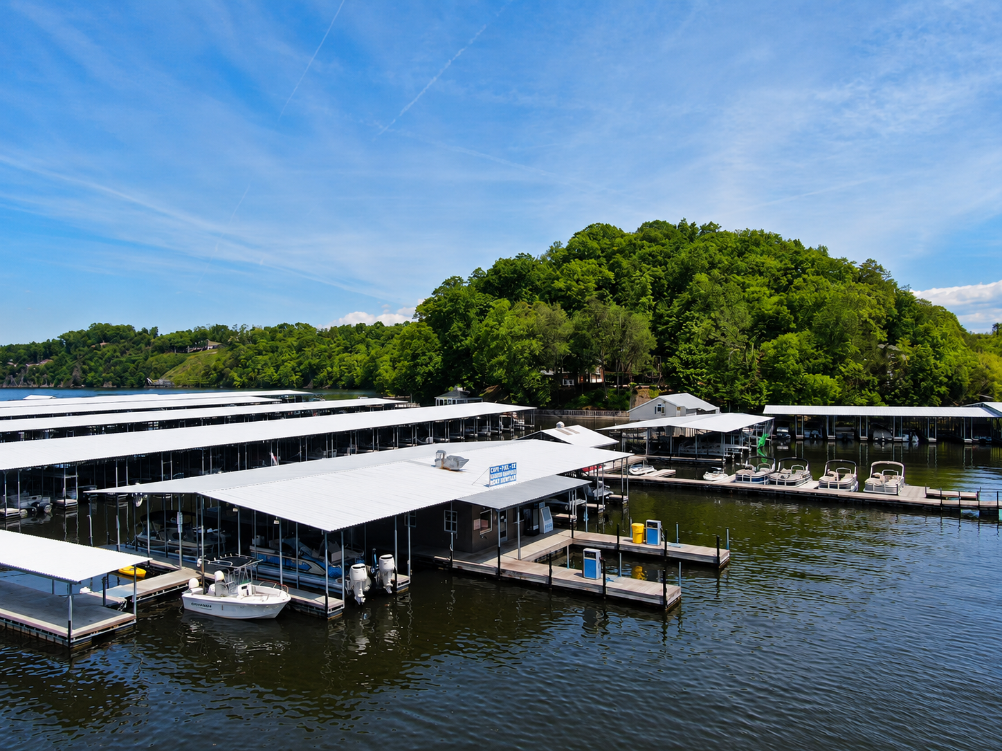 Covered marina docks beside calm water, with boats and a tree-covered shoreline under a blue sky.