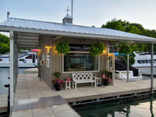 Explore More About What We Do! Small waterfront dockside kiosk with hanging plants, white benches, and boats in the background