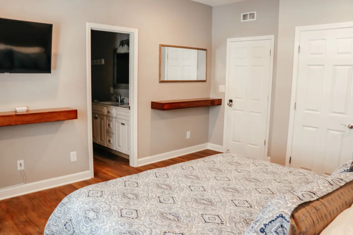 Bedroom with beige walls, white doors, a wall-mounted TV, and a bathroom doorway.