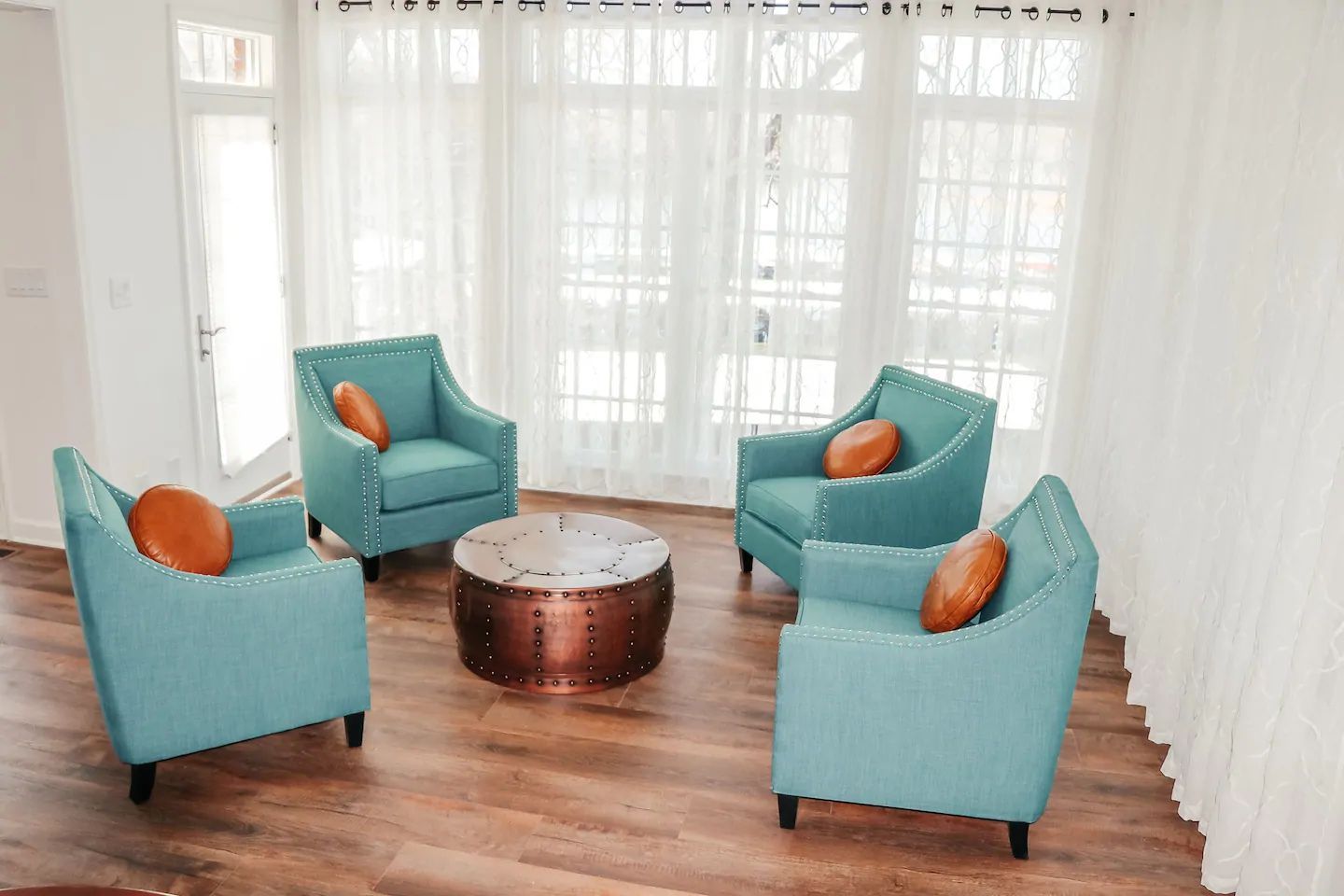 Turquoise chairs with orange pillows arranged around a round coffee table in a bright room with sheer curtains