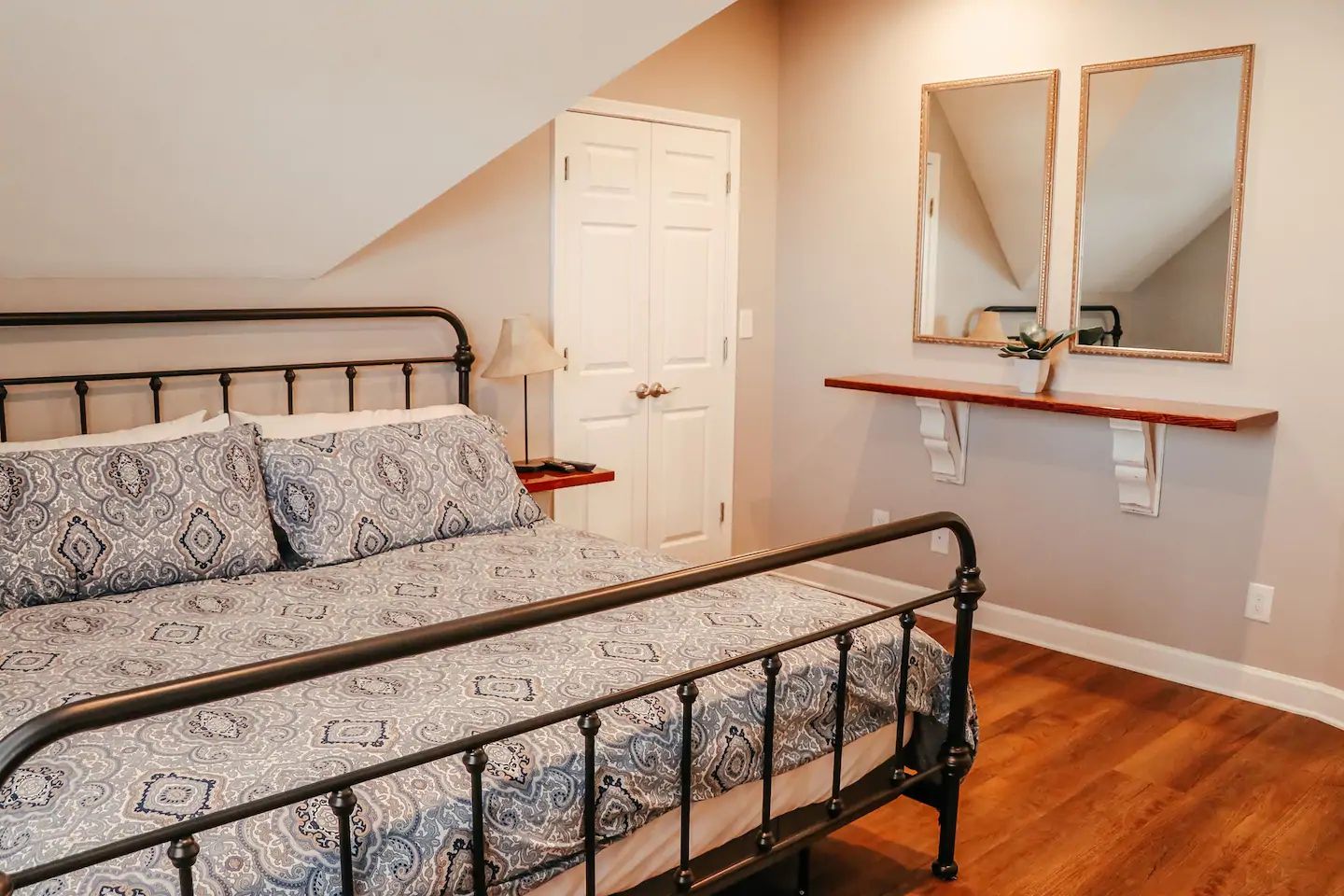 Bedroom with black metal bed, patterned bedding, hardwood floor, and wall mirror above a small shelf.