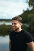 Smiling man in a black T-shirt standing outdoors near a lake and trees