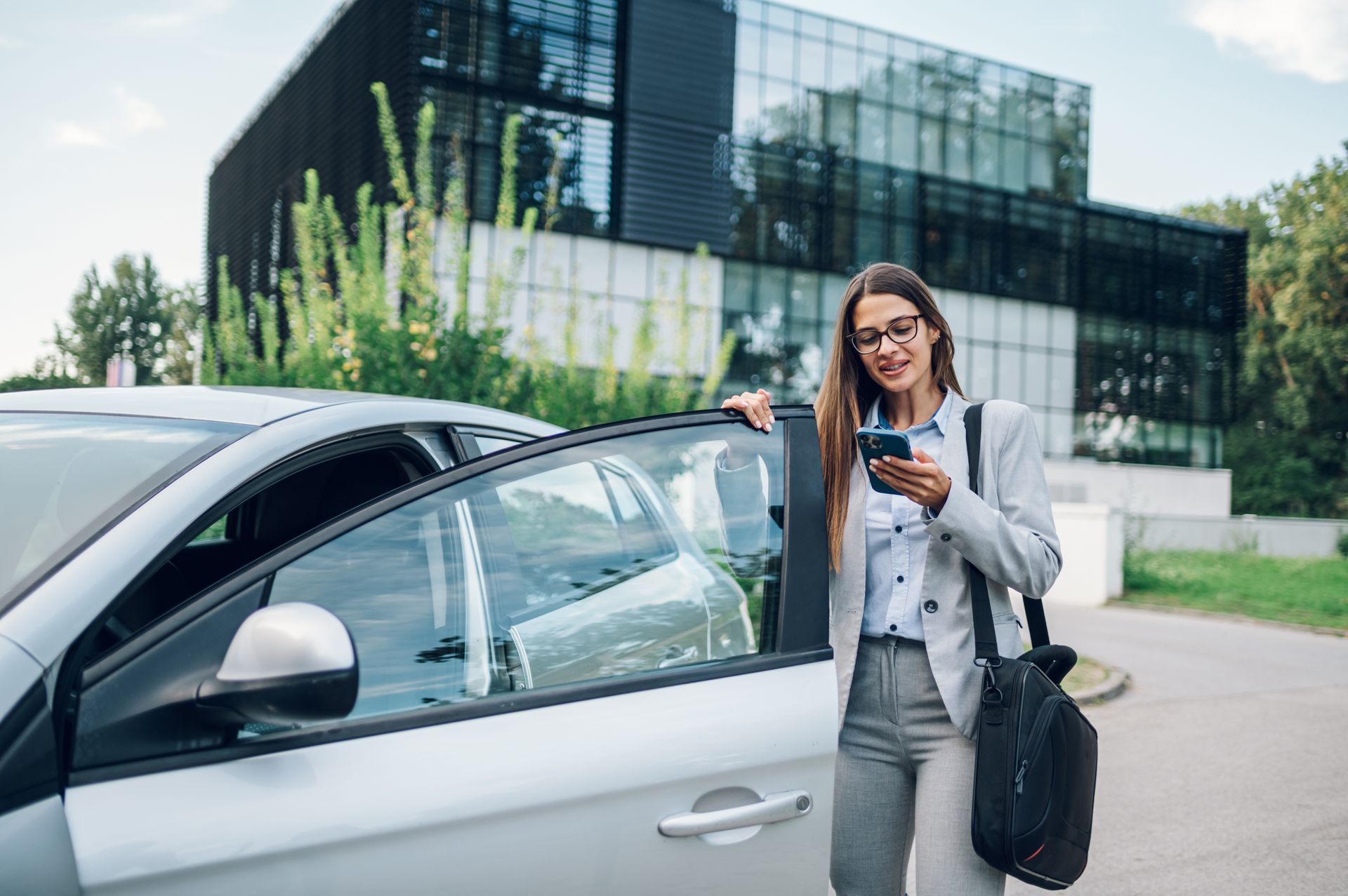 Woman in business attire using a phone next to a car, in front of a modern building.