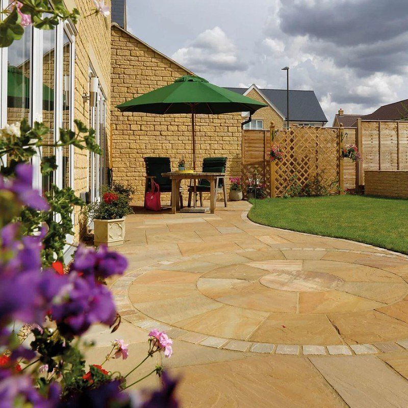 A patio with a circular stone design, a green umbrella, table and chairs, next to a stone house and lawn under a cloudy sky.