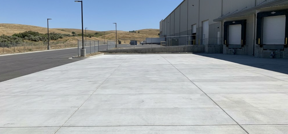 A wide, clean commercial concrete driveway leading to the loading docks of a large commercial warehouse, with dry grassy hills in the background.