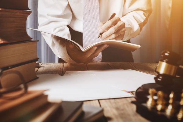 Person writing in a notebook at a desk with law books, a gavel, and a blank paper.