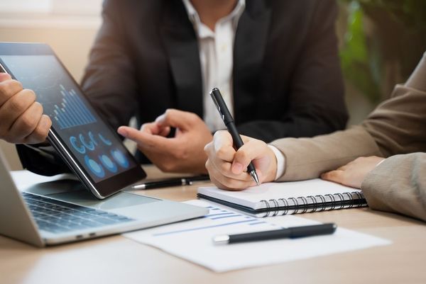 People in business attire reviewing data on a tablet, taking notes at a desk.