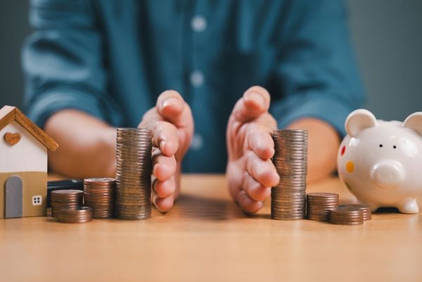 Person's hands framing stacks of coins, with house model and piggy bank, suggesting financial planning.