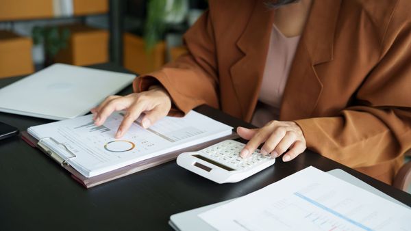 Person in brown blazer using a calculator and reviewing financial reports at a desk with a laptop and boxes.