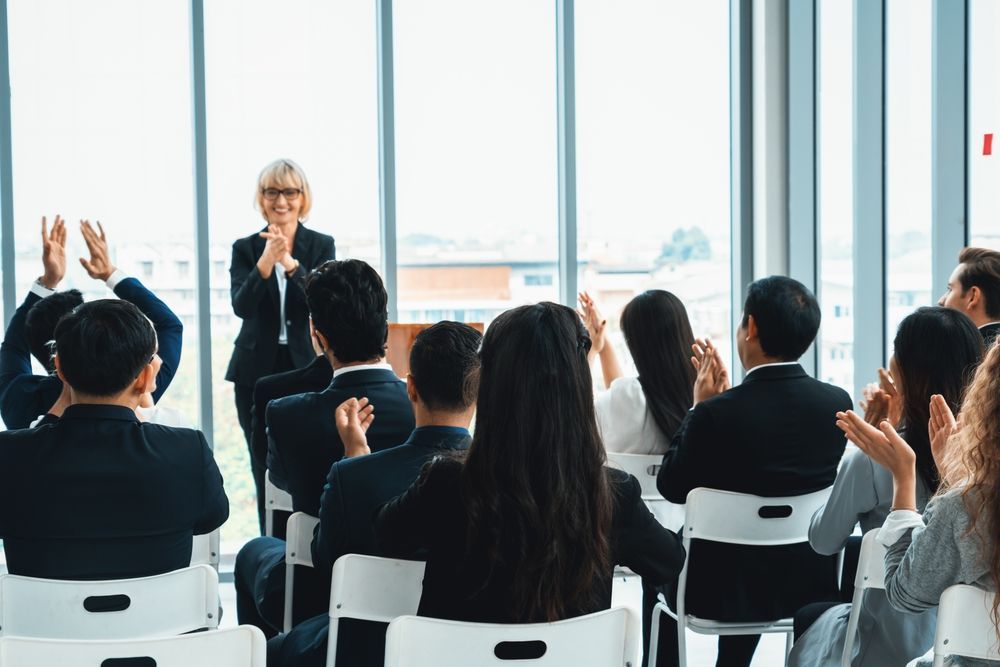 A woman in a suit speaks at a podium as audience applauds in a bright, modern room.