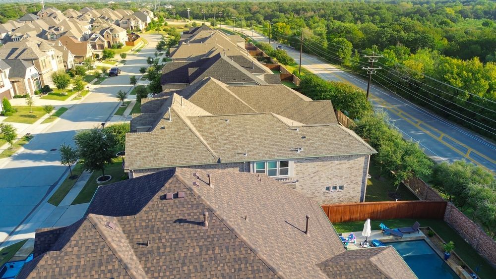 Aerial view of suburban homes with brown roofs, a road, and green trees.