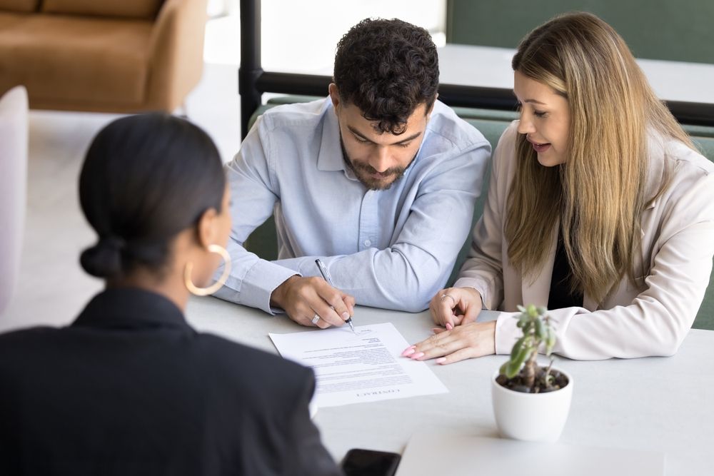Woman in blazer with couple signing documents at table, small plant.