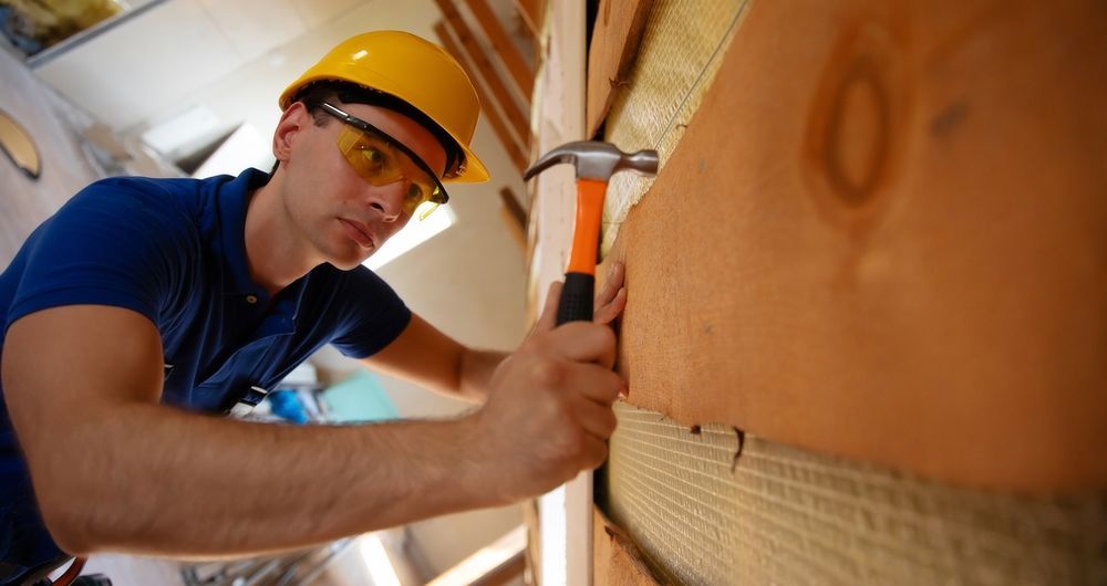 Construction worker hammering insulation in an attic, wearing a yellow hard hat and safety glasses.