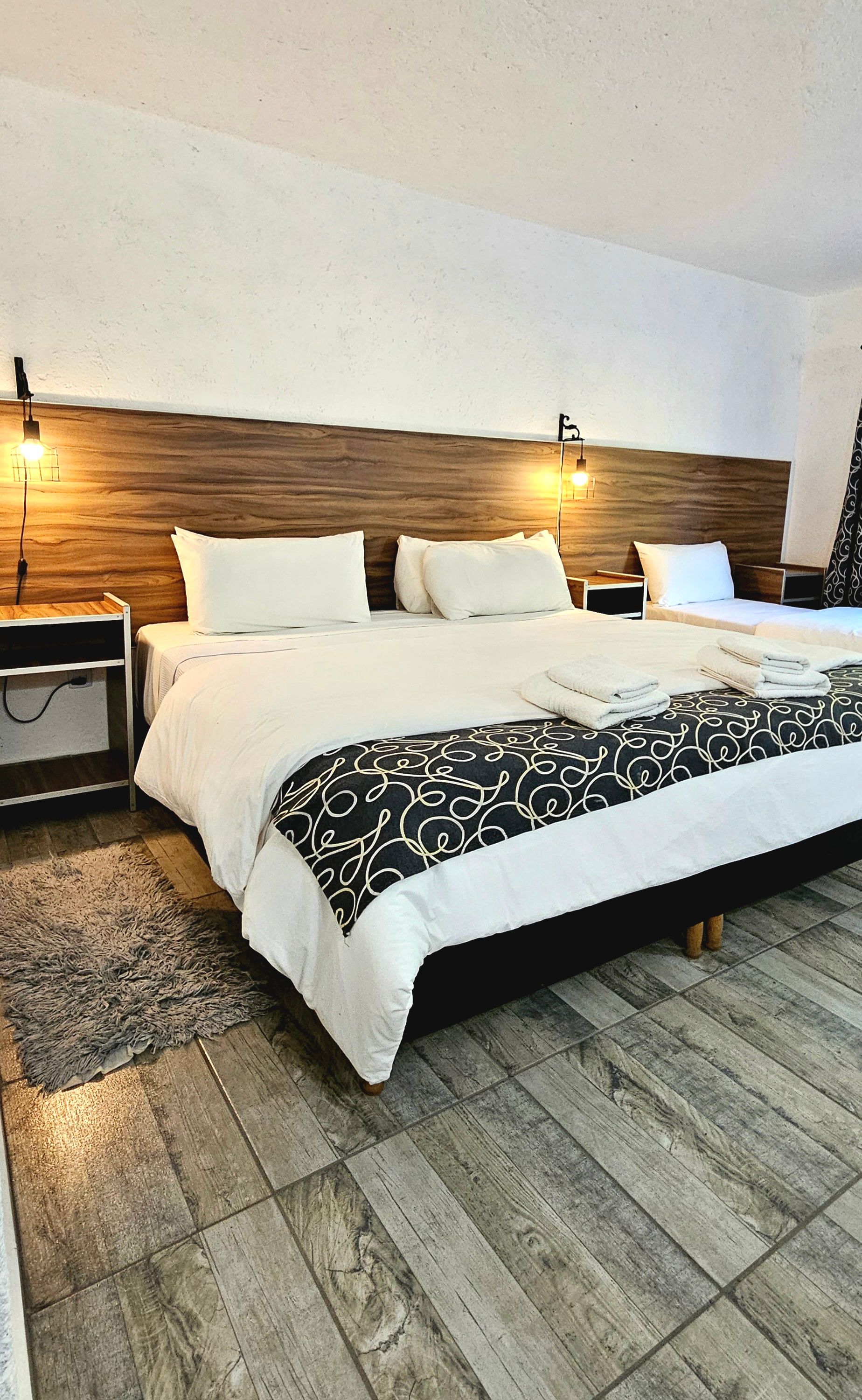 Bedroom with a large bed and smaller bed.  White bedding, black patterned blanket, wooden headboard, and wood-look flooring.
