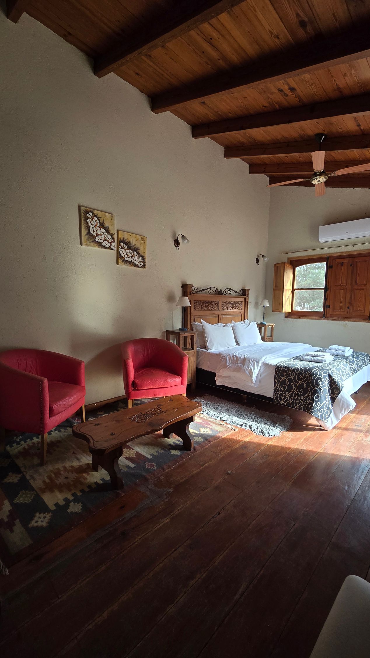 Bedroom with wood ceiling, bed, two red chairs, and wooden table on a wooden floor.