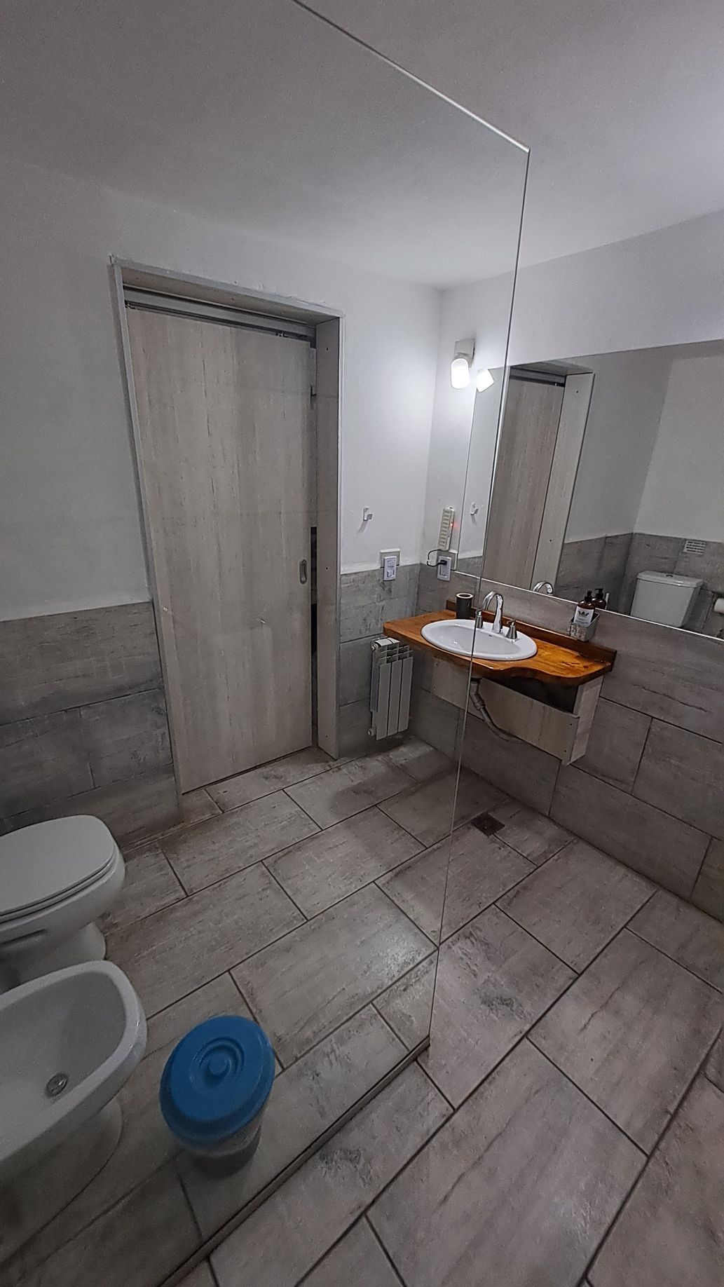 Bathroom with white tile floors, white toilet and bidet.  Sink on a wooden shelf, white door.