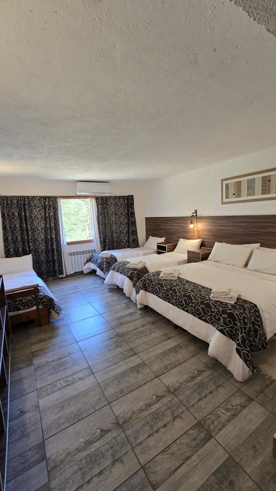 Spacious hotel room with four beds, wood-look flooring, patterned curtains, and a window.