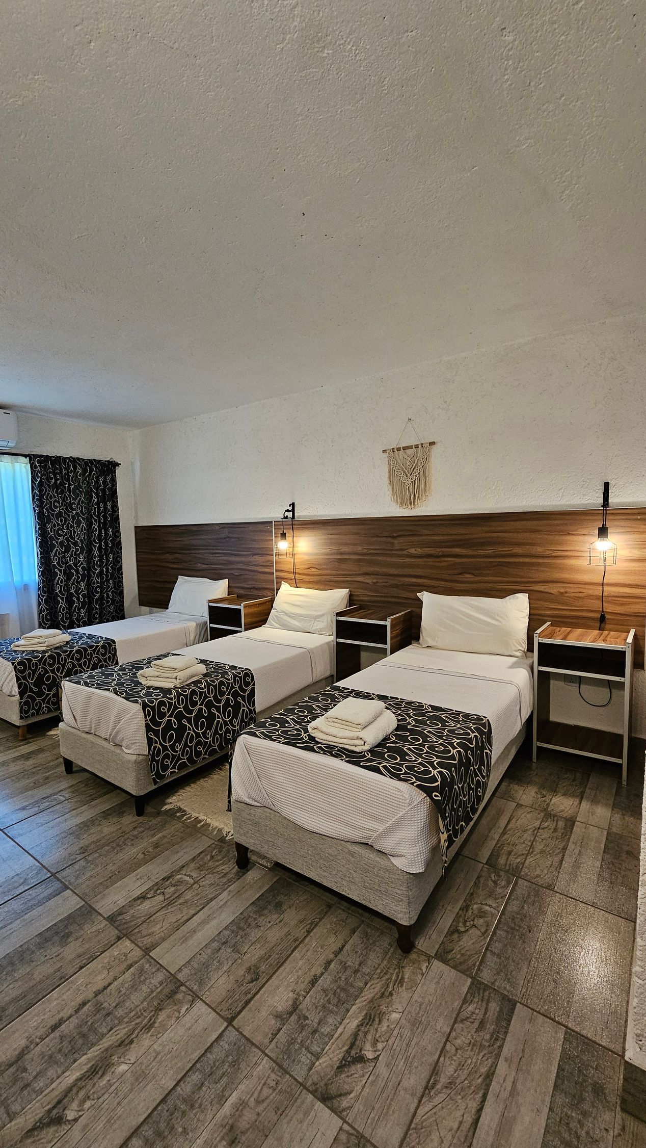 A hotel room with three single beds, wooden headboard, bedside tables, and patterned bedding.