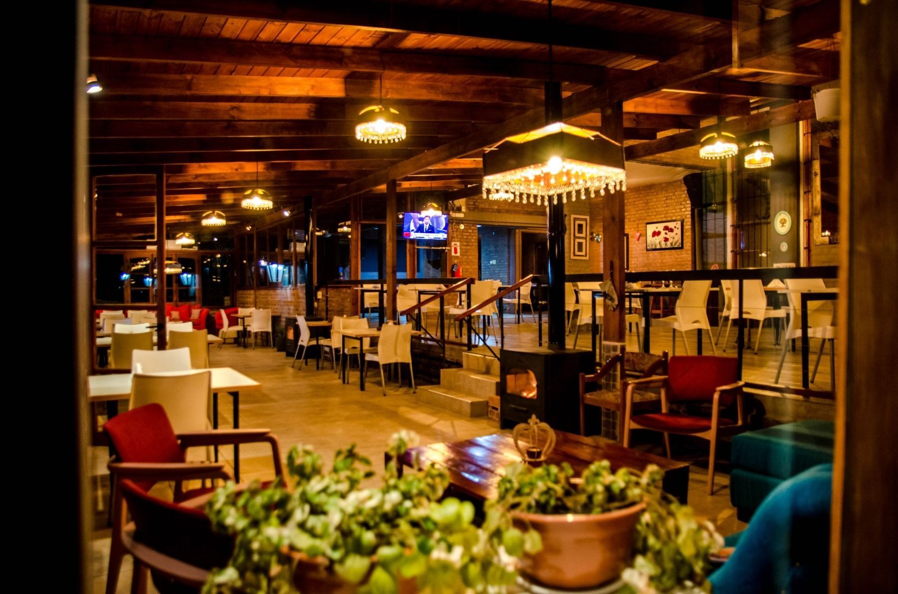 Restaurant interior with tables, chairs, and hanging lights; wooden beams and warm lighting create a cozy atmosphere.