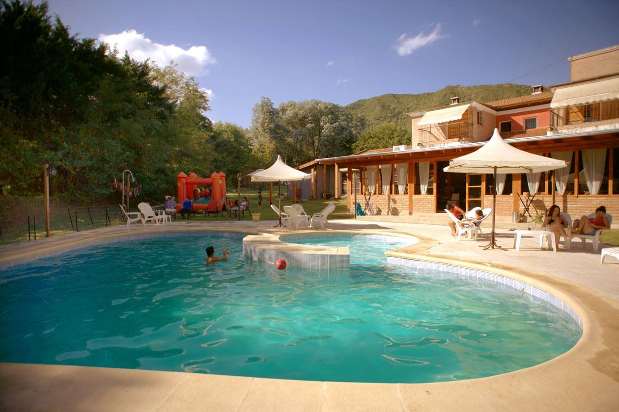 Pool with turquoise water, lounge chairs, and people near a lodge with a mountain backdrop.