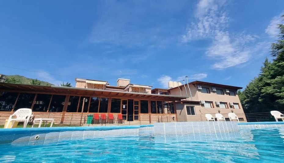 Pool with building in the background. Blue water and sky. Wooden porch, trees and white chairs.