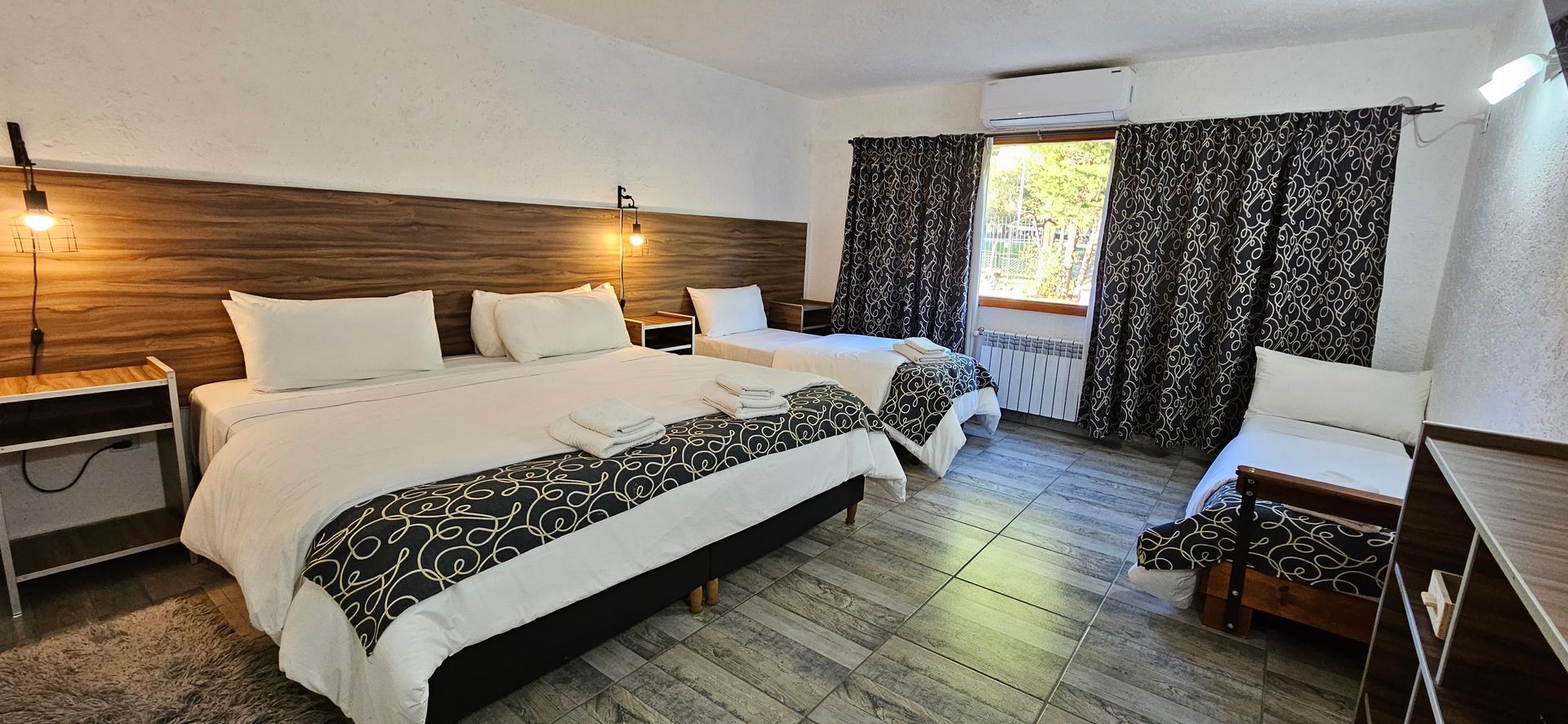 Hotel room with three beds, wooden headboards, patterned bedspreads, and dark curtains.