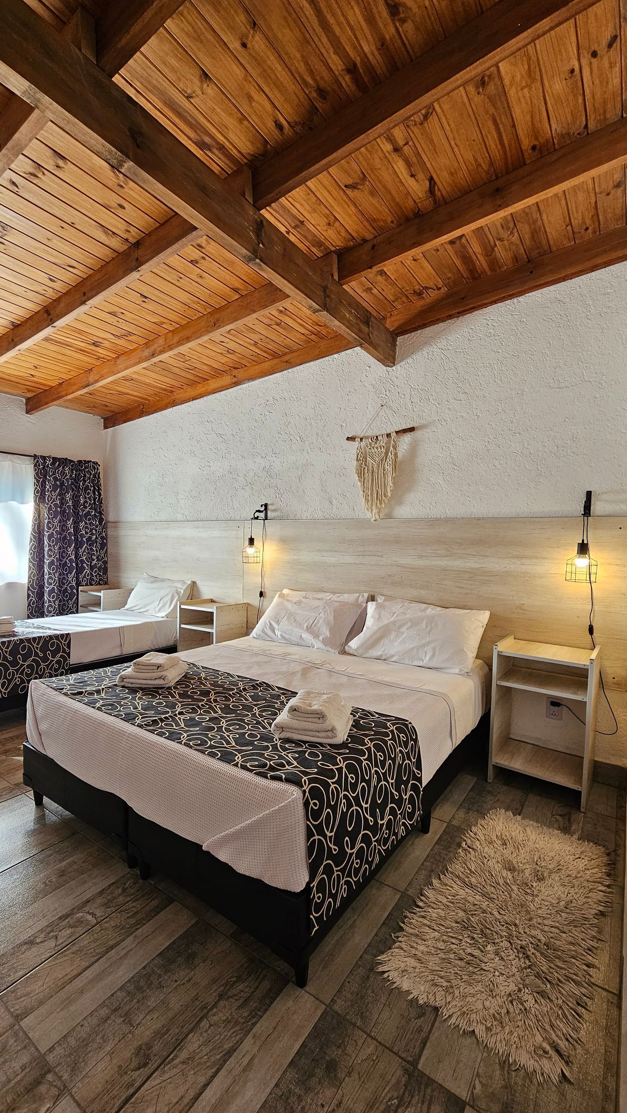 Bedroom with wood ceiling, large bed with dark patterned cover, single bed, and textured white wall.
