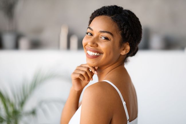 A woman in a white tank top is smiling and looking over her shoulder.