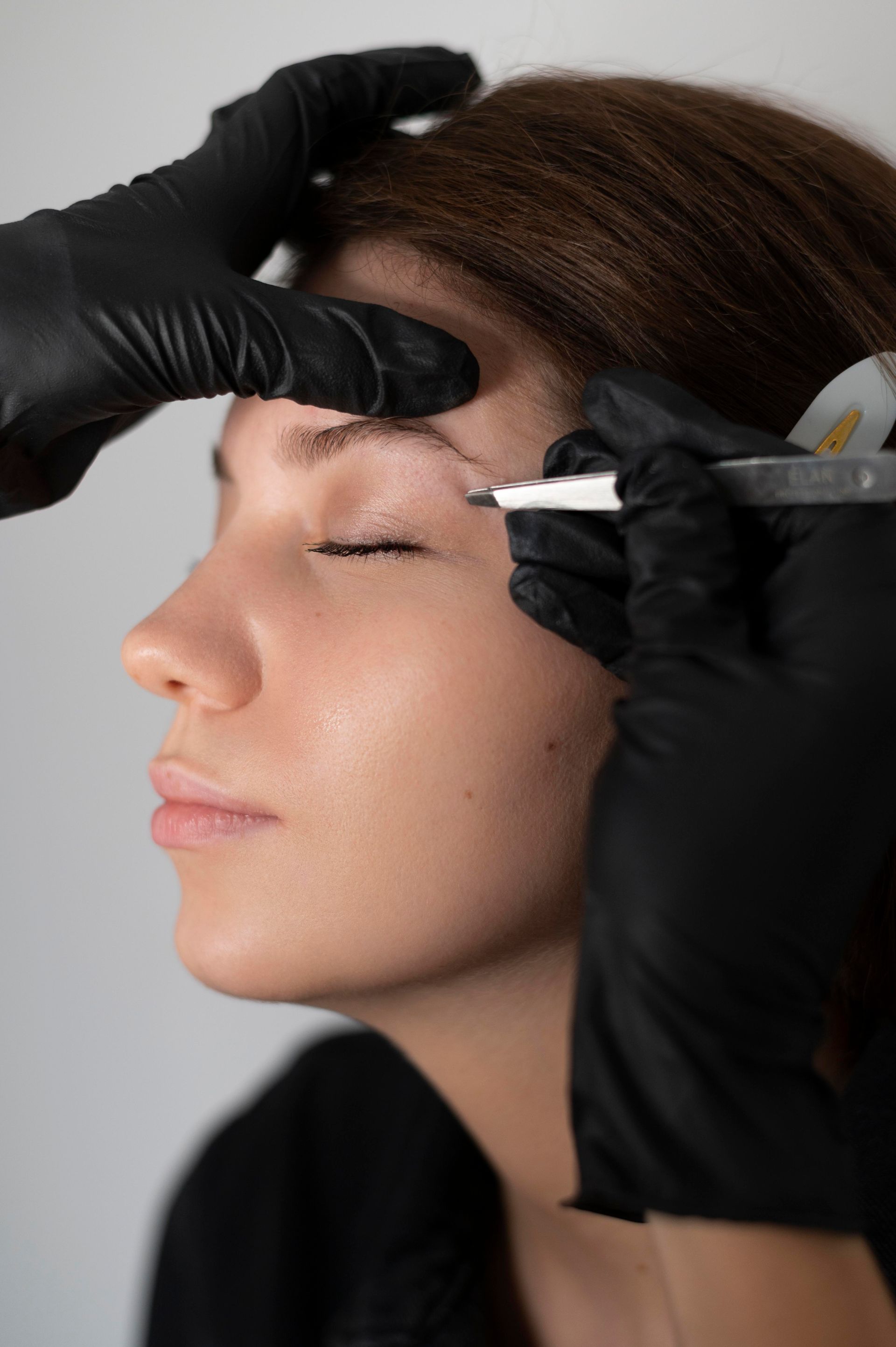 A woman is getting her eyebrows done by a person wearing black gloves.