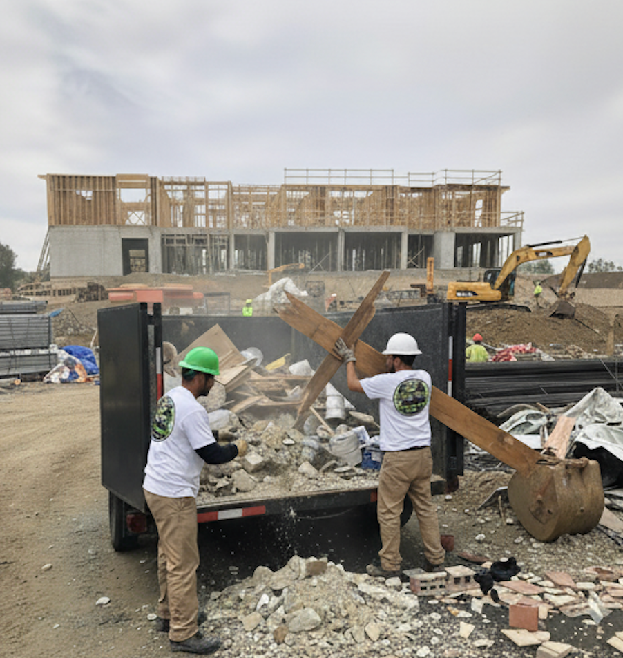 Two workers loading debris into a truck at a construction site. Wooden structure under construction is in the background.