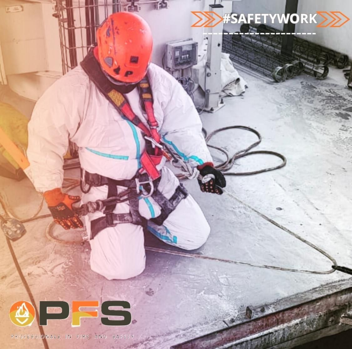 Un trabajador con traje de materiales peligrosos y casco de seguridad arrodillado sobre una superficie metálica, con un arnés de seguridad colocado.