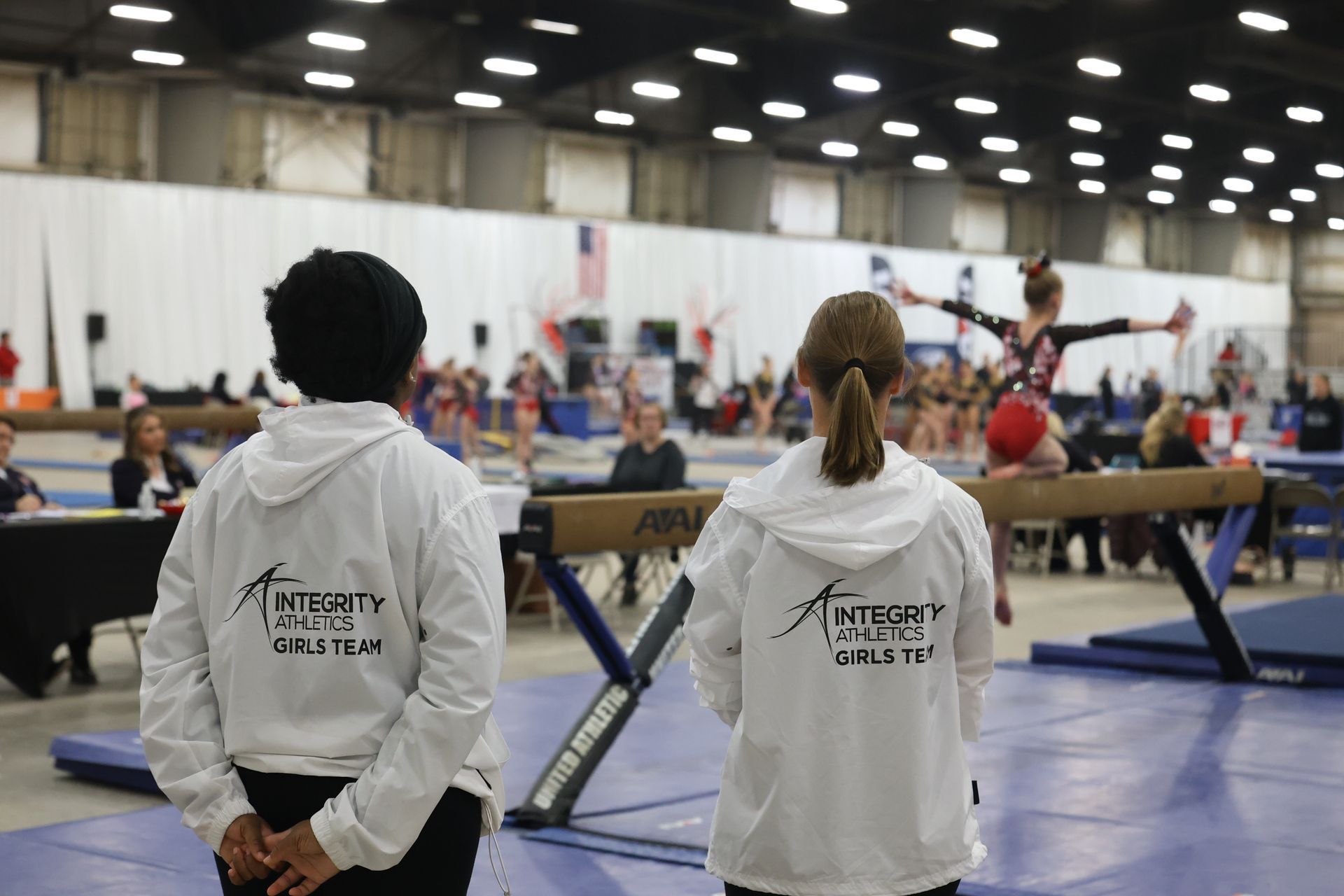 A man and a woman are standing in front of a gymnast on a balance beam.