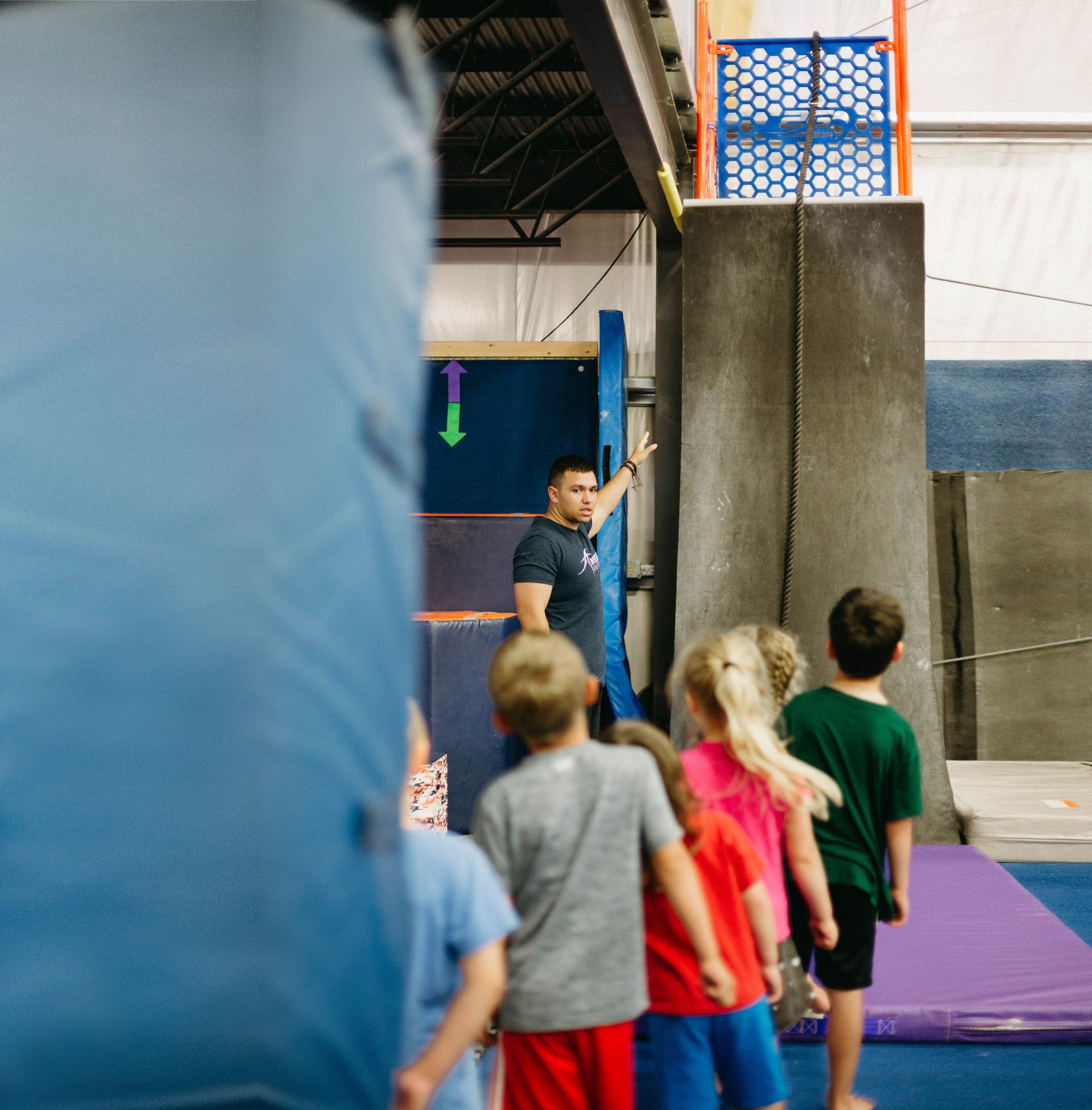 A group of children are standing in a gym watching a man climb a ladder