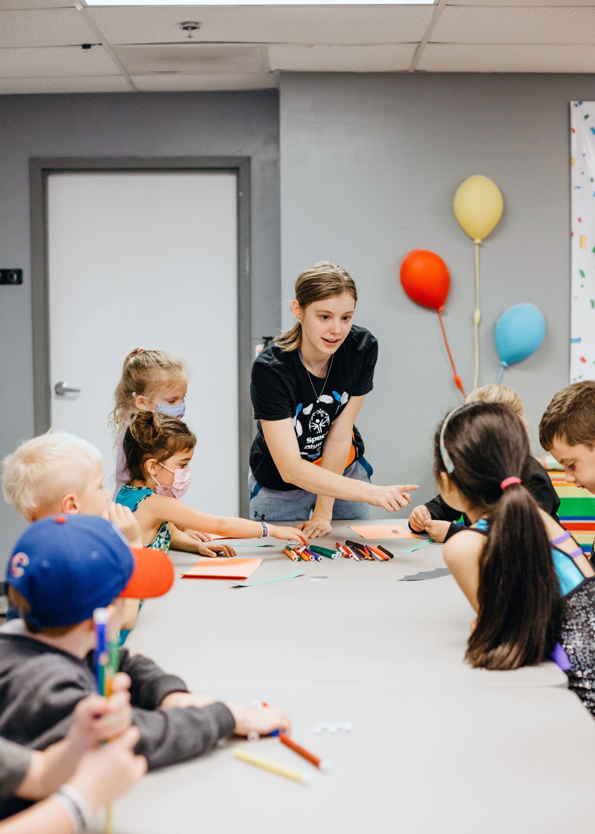 A group of children are sitting around a table with balloons on the wall.
