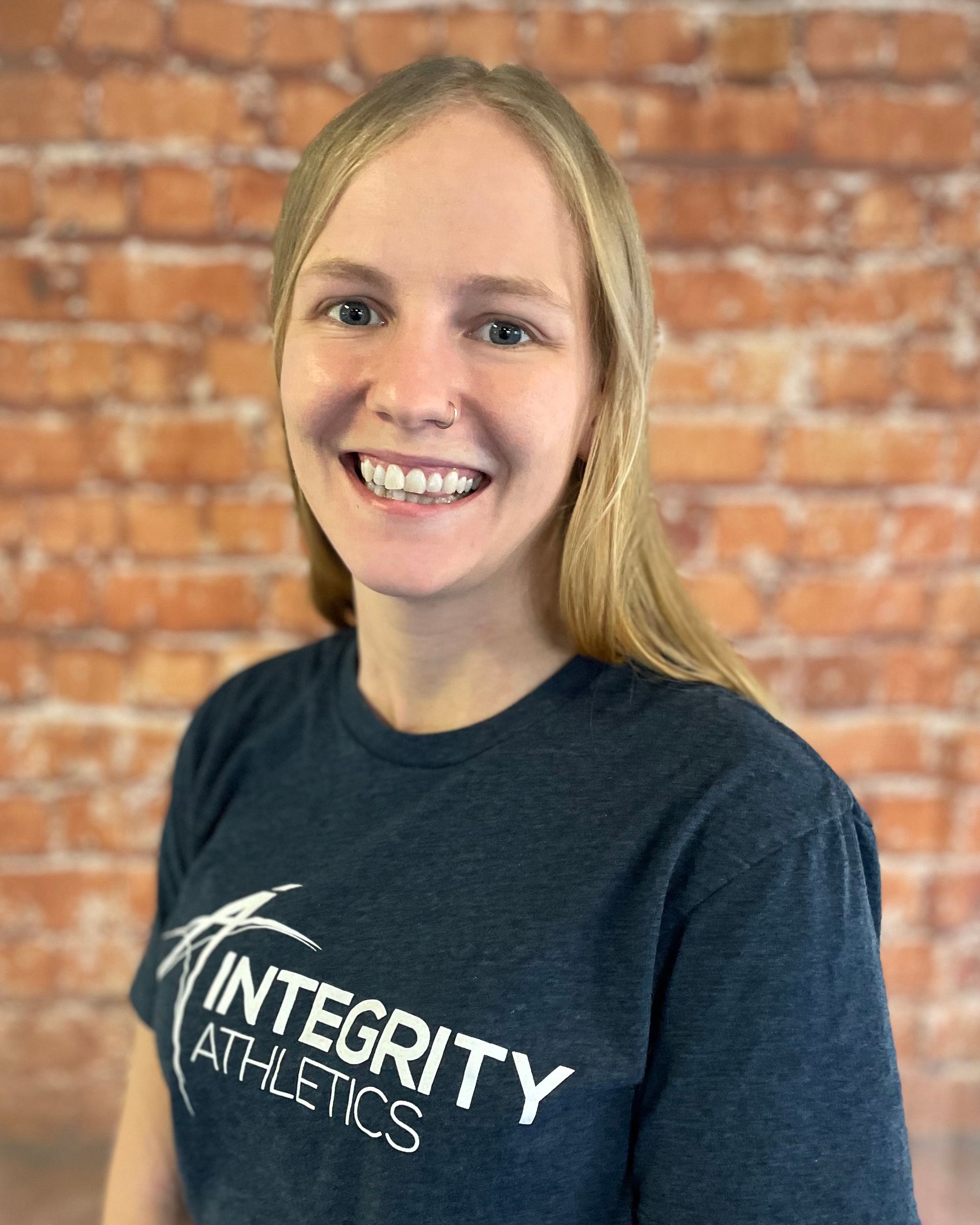 A woman wearing a t-shirt that says integrity athletics is smiling in front of a brick wall.