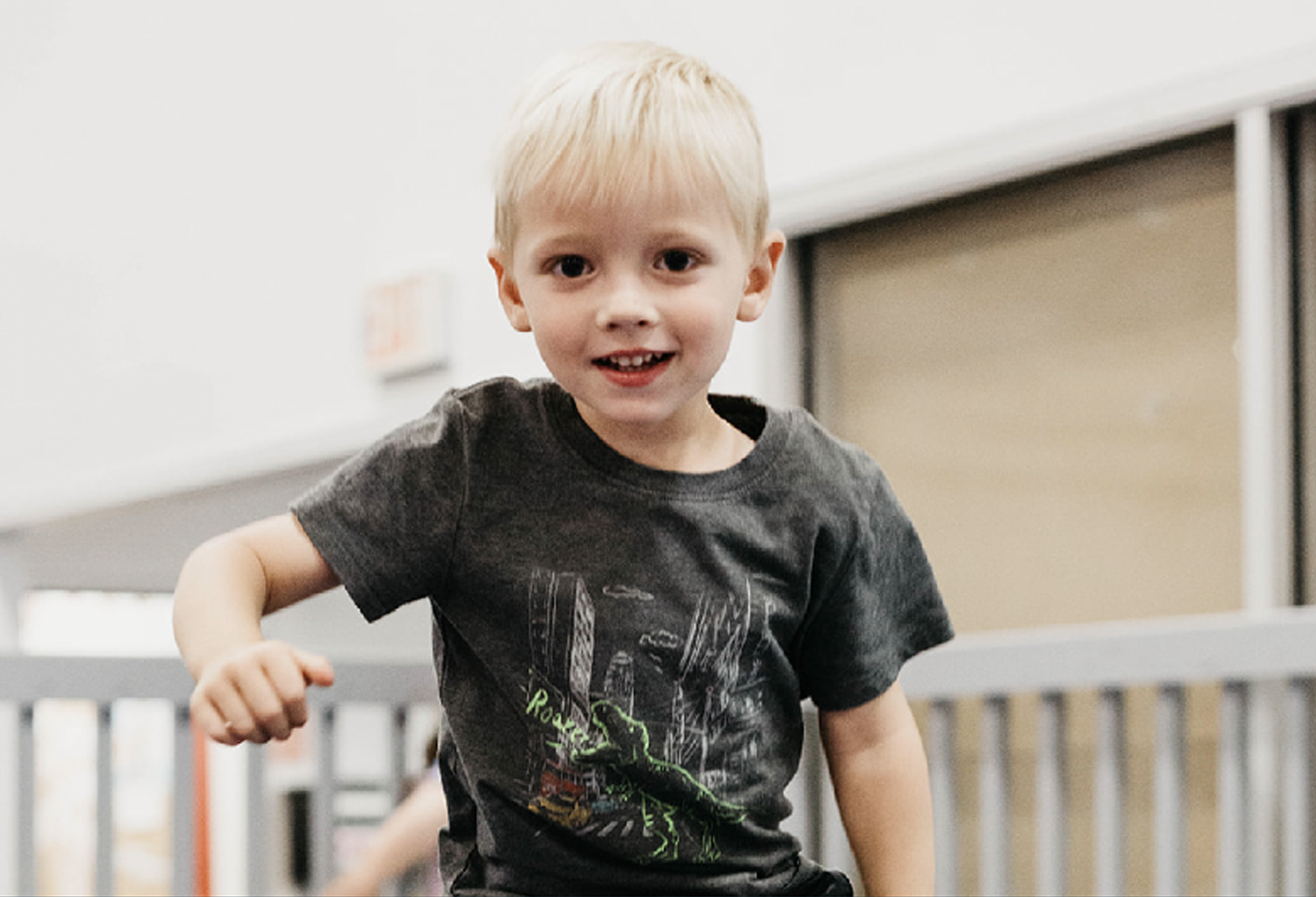 A young boy in a gray shirt is standing on a trampoline and smiling.