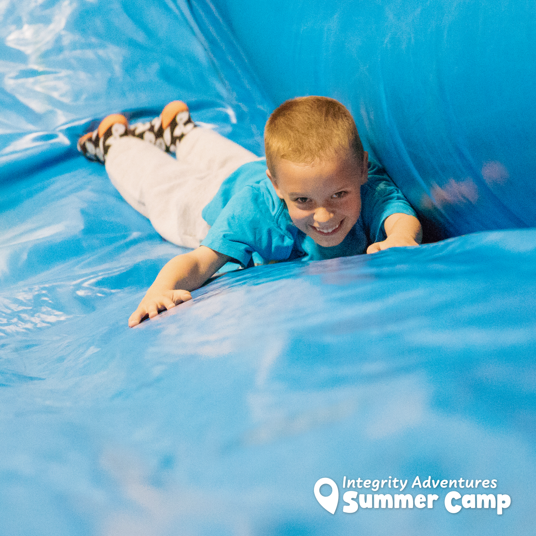 A young boy is sliding down a blue inflatable slide sponsored by integrity adventures summer camp