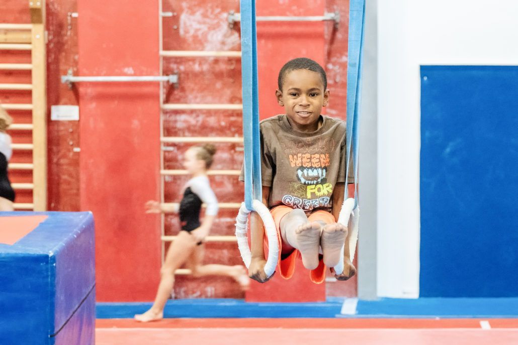 A young boy is sitting on a pair of gymnastic rings in a gym.
